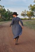 Person in a blue navy check dress and hat walking on a dirt road with trees in the background
