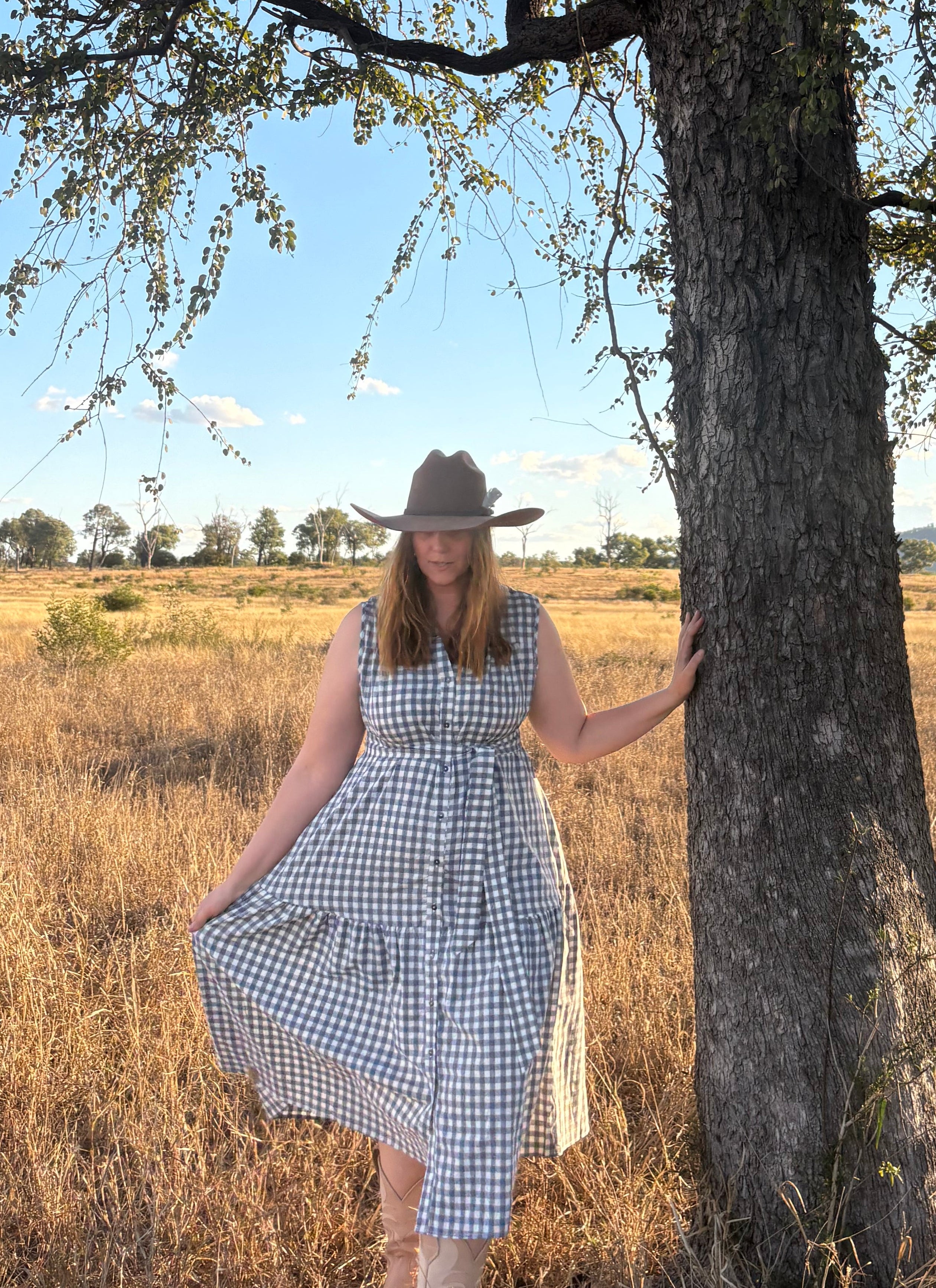 A woman wearing a blue and white gingham dress and a cowboy hat stands next to a large tree in a dry grassy field under a blue sky.