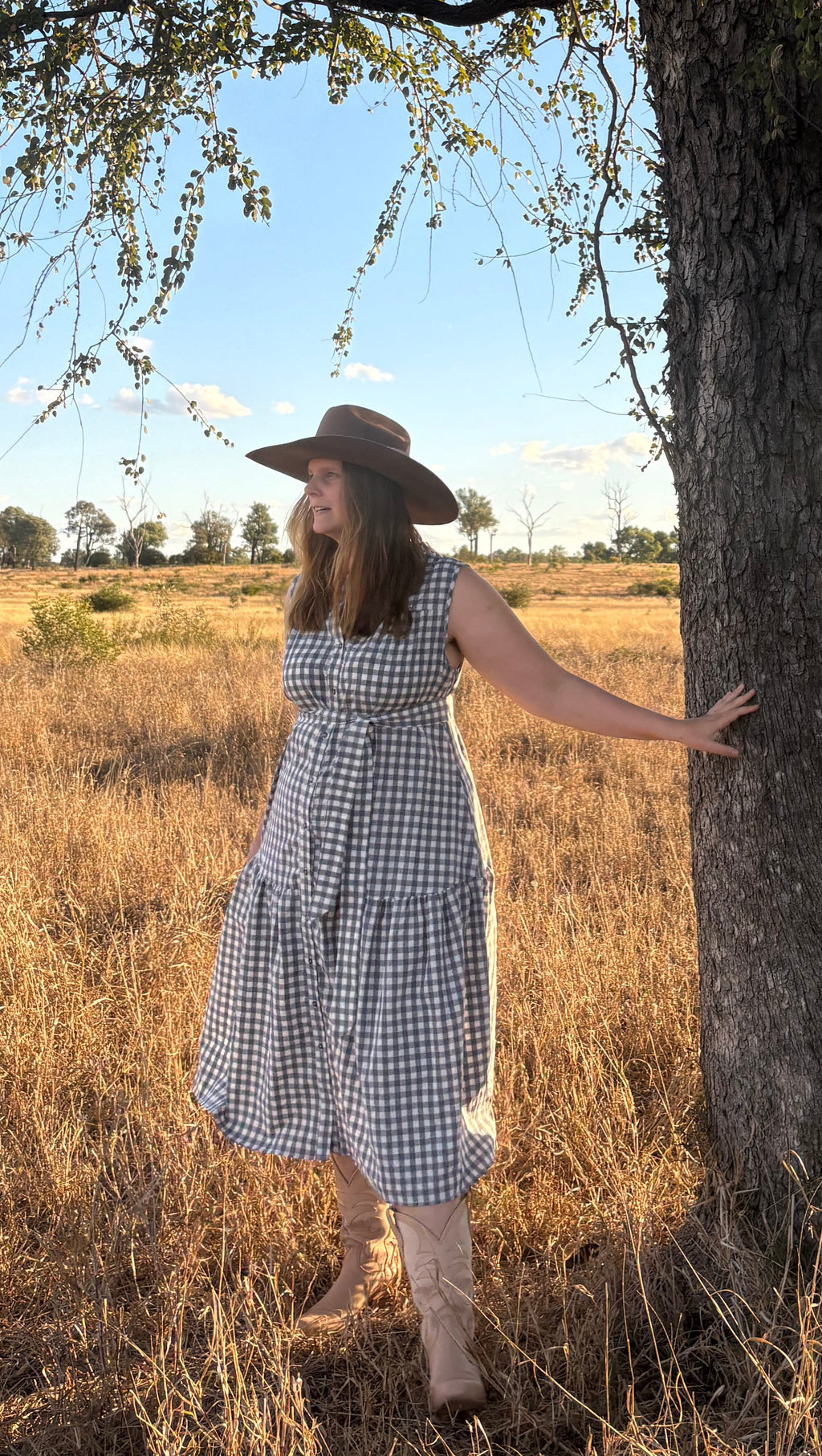 A woman wearing a blue and white gingham midi dress and a wide-brimmed brown hat stands in a dry grassy field next to a large tree under a blue sky.