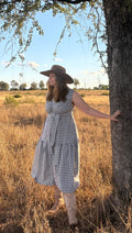 A woman wearing a blue and white gingham midi dress and a wide-brimmed brown hat stands in a dry grassy field next to a large tree under a blue sky.