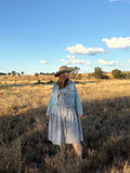 A woman stands in a dry grassy field under a bright blue sky with scattered clouds, wearing a wide-brim straw hat, a light blue denim jacket, and a blue-and-white gingham dress.